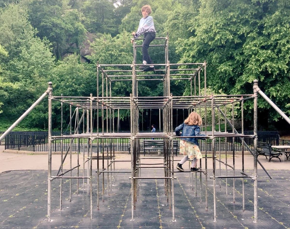 Two children are playing on a large metal climbing structure in a park, surrounded by trees. One child stands at the top, while the other is climbing up from the side. The scene is outdoors on a cloudy day.