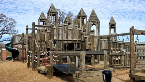 A large wooden playground structure resembling a castle with multiple towers, bridges, slides, and climbing areas, set on a gravel surface under a partly cloudy sky.