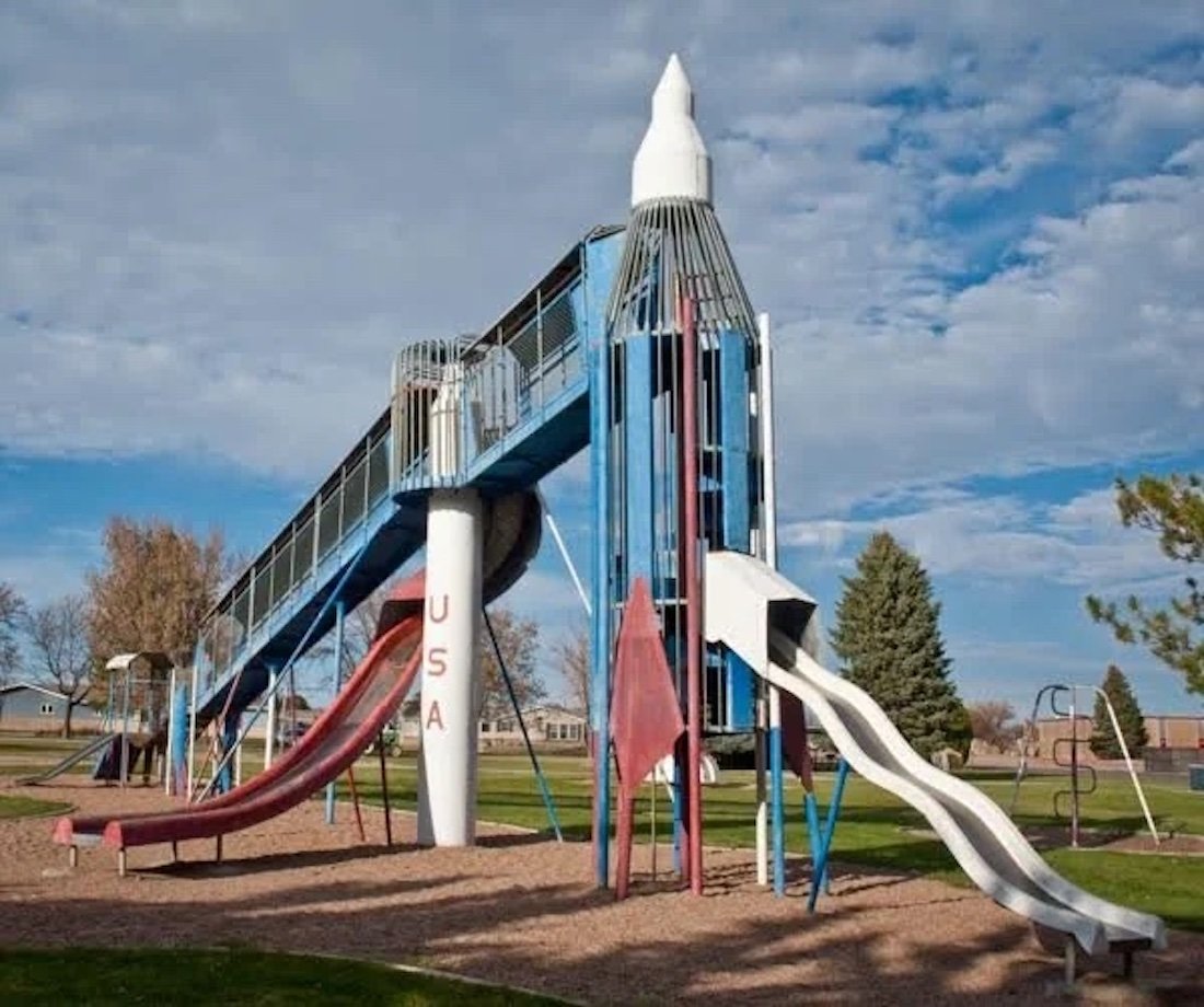 A playground slide shaped like a large rocket ship with "USA" written on its side, featuring metal bars, ladders, and slides. The structure stands on mulch with trees and a blue sky in the background.