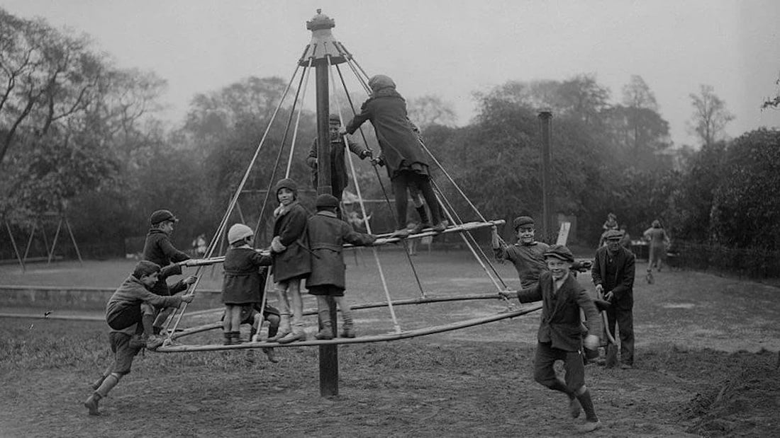 Black and white photo of children in coats and hats playing on a large, pyramid-shaped metal climbing frame in a park, with trees and a swing set in the background.