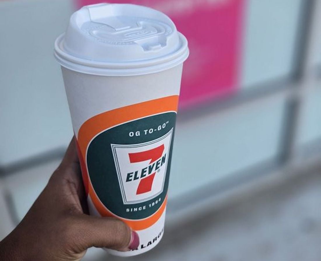 A hand holding a large white 7-Eleven coffee cup with a plastic lid, featuring the store's logo and orange-green design. The background is blurred with shades of pink and gray.