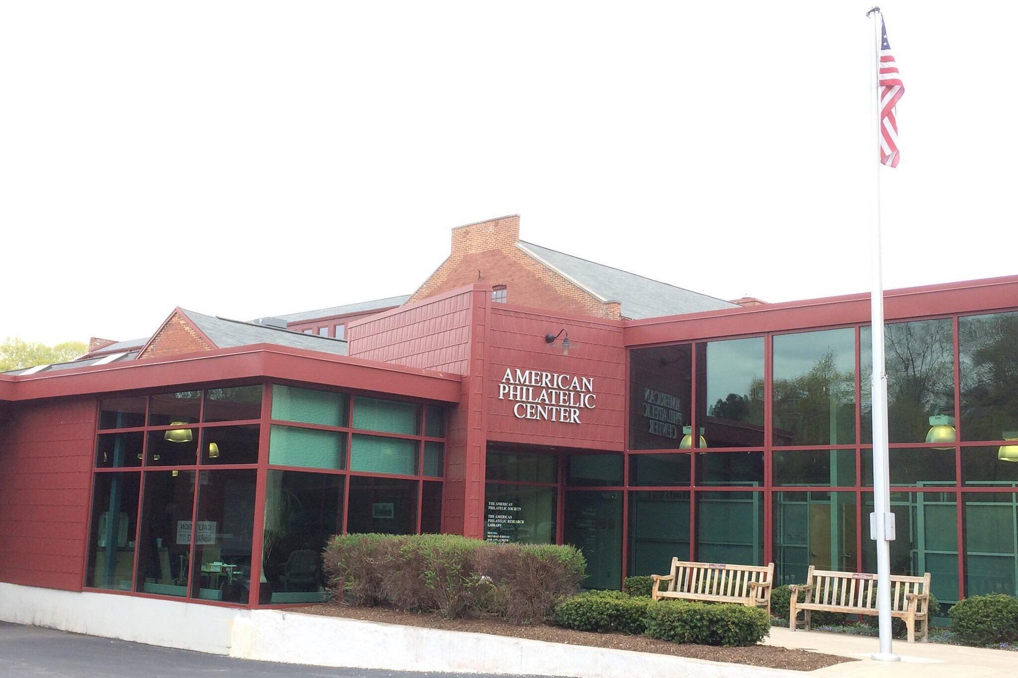 Front exterior of American Philatelic Center, Bellefonte, Pennsylvania, lots of windows and dark red color, bushes with seating and an American flag outside