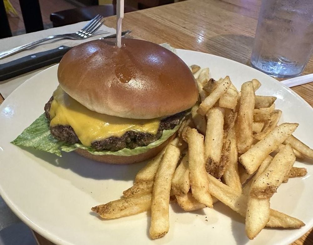 A plate with a cheeseburger topped with lettuce and a slice of cheddar cheese in a shiny bun, served alongside a pile of golden French fries. A fork, knife, and glass of water are visible in the background.