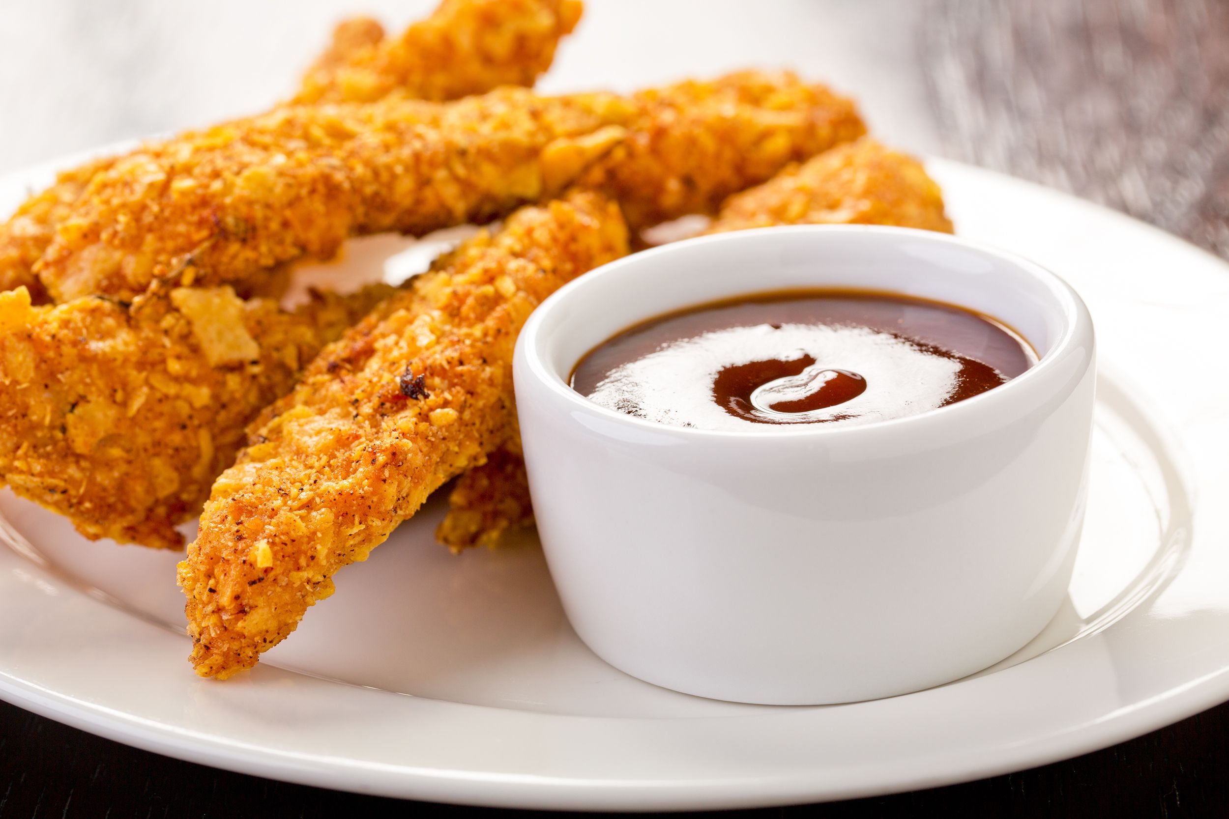 Baked chicken fingers on a white plate with BBQ dipping sauce in a white cup with a blurred background of a wooden table