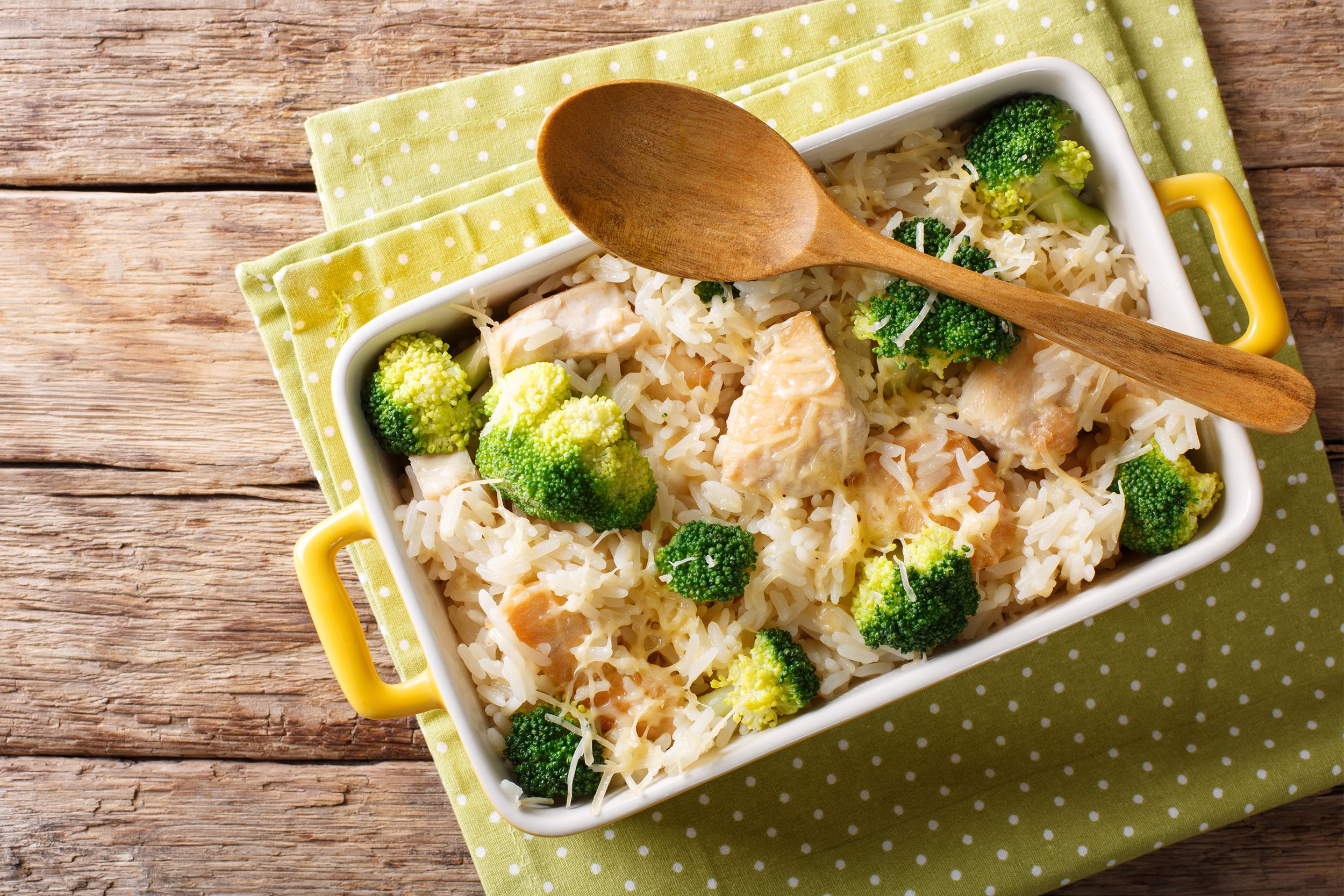 Broccoli pesto chicken & rice in a yellow rectangle ceramic cooking pot with a wooden serving spoon on top on green polka dot napkins on a rustic wooden table