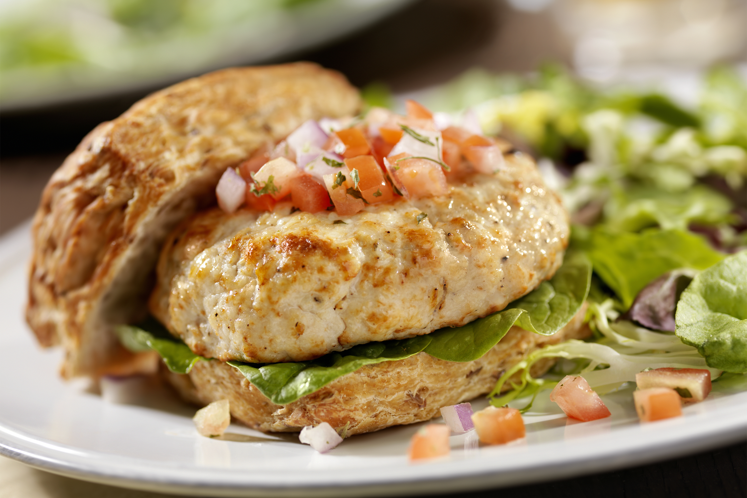 Closeup of a chicken burger on a whole wheat bun with a diced tomato topping on a white plate with a blurred background of lettuce