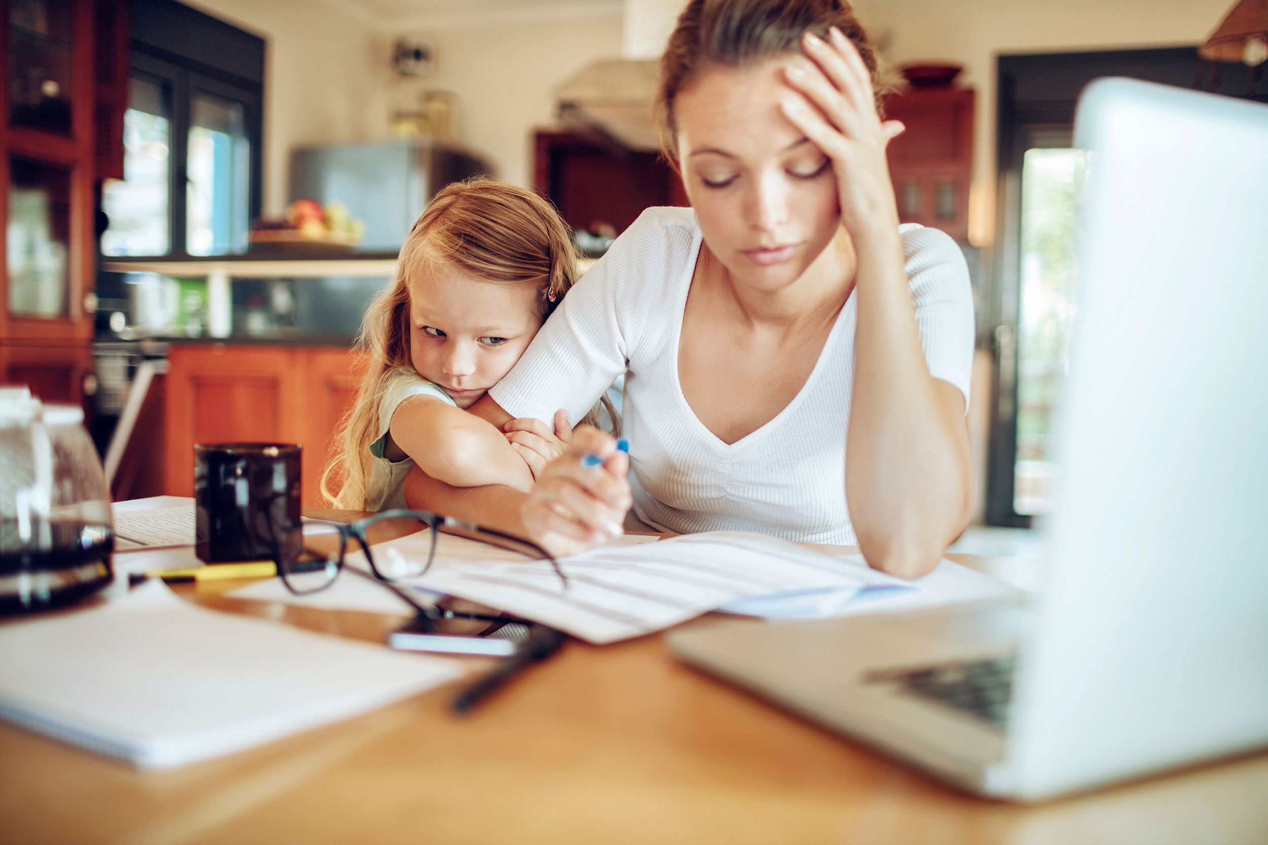 Closeup of stressed young mother at desk with laptop and scattered papers in foreground, worried little girl is hugging her arm, with a blurred background of kitchen and living room of home