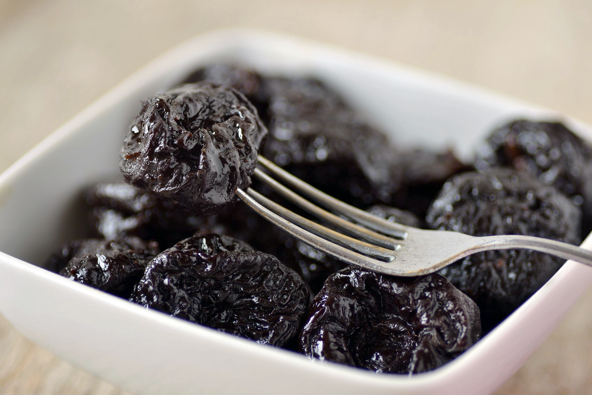 Closeup of prunes in a square white porcelain bowl, with a fork over them, into one prune, selective focus, on a beige table