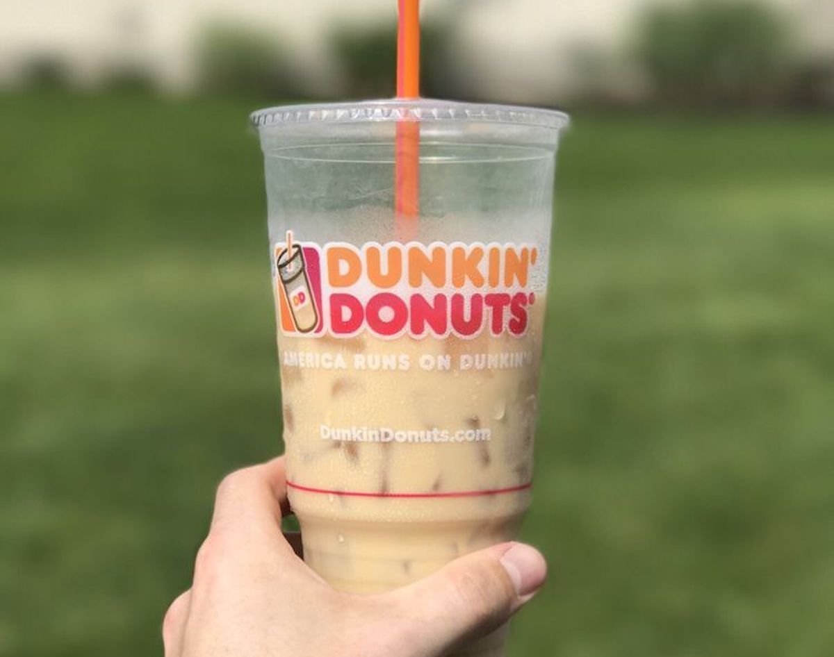 A hand holds a clear plastic Dunkin’ Donuts cup filled with iced coffee and an orange straw, with a blurred grassy background.