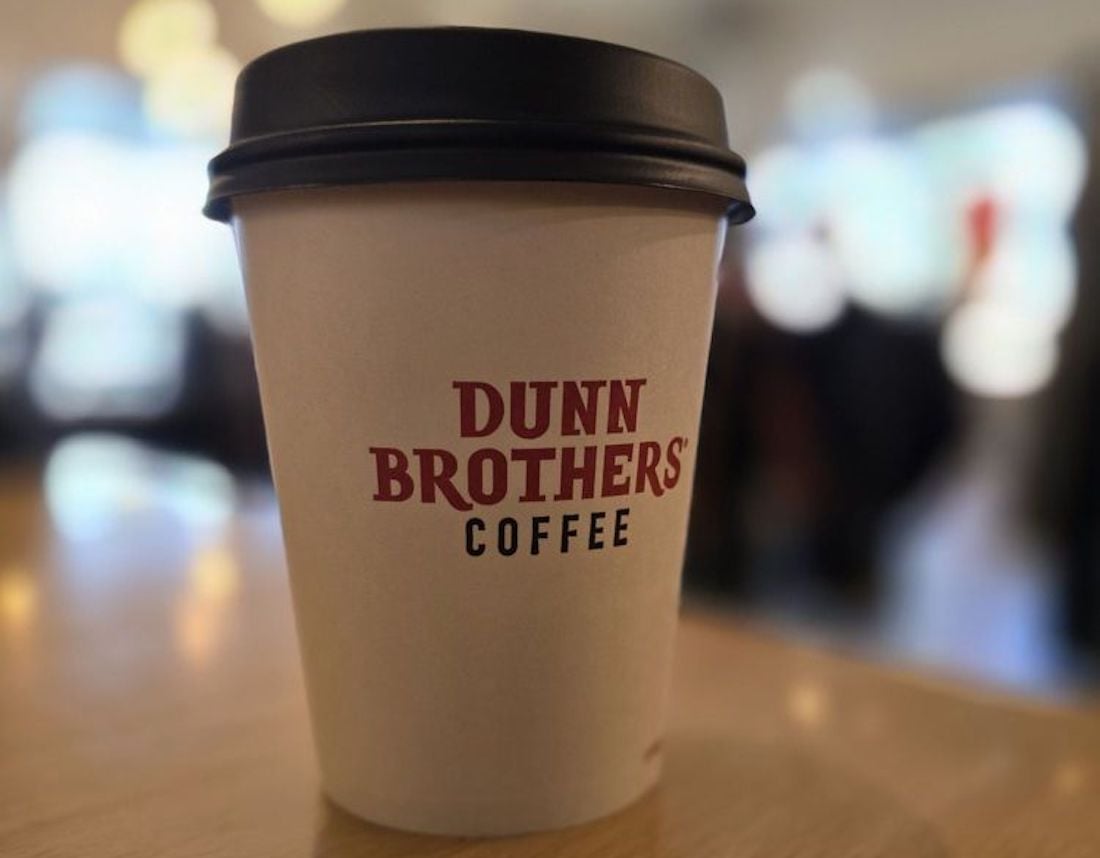 A white disposable coffee cup with a black lid sits on a table. The cup features the red logo "Dunn Brothers Coffee." The background is blurred, suggesting a café environment.