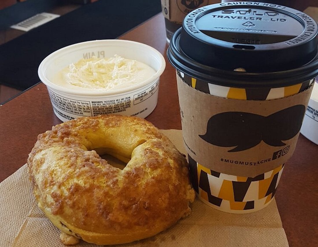 A cinnamon crumb bagel on a napkin, a cup of coffee with a sleeve featuring a mustache design, and a container of plain cream cheese on a table.