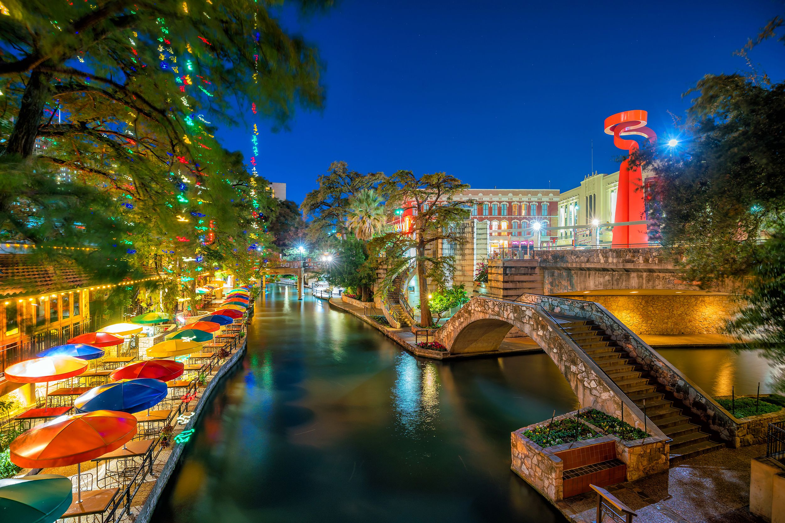 Empty River Walk, San Antonio lit up during late evening, colorful umbrellas on tables on the left, river in the middle, and brick walkway on the right
