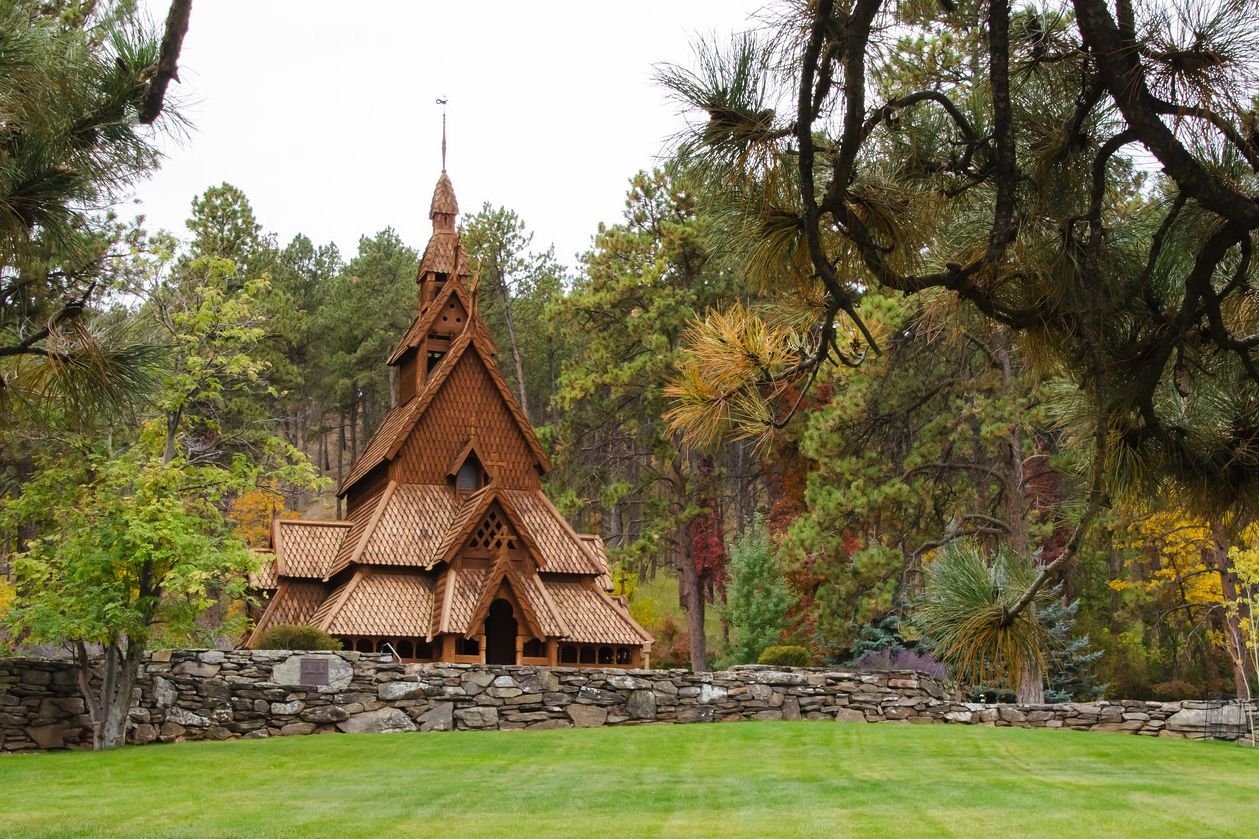 Exterior of Chapel in the Hills, Rapid City, South Dakota, flat stone fence, grass in the foreground, surrounded by trees and plants, a tree in the foreground on both left and right, during summer