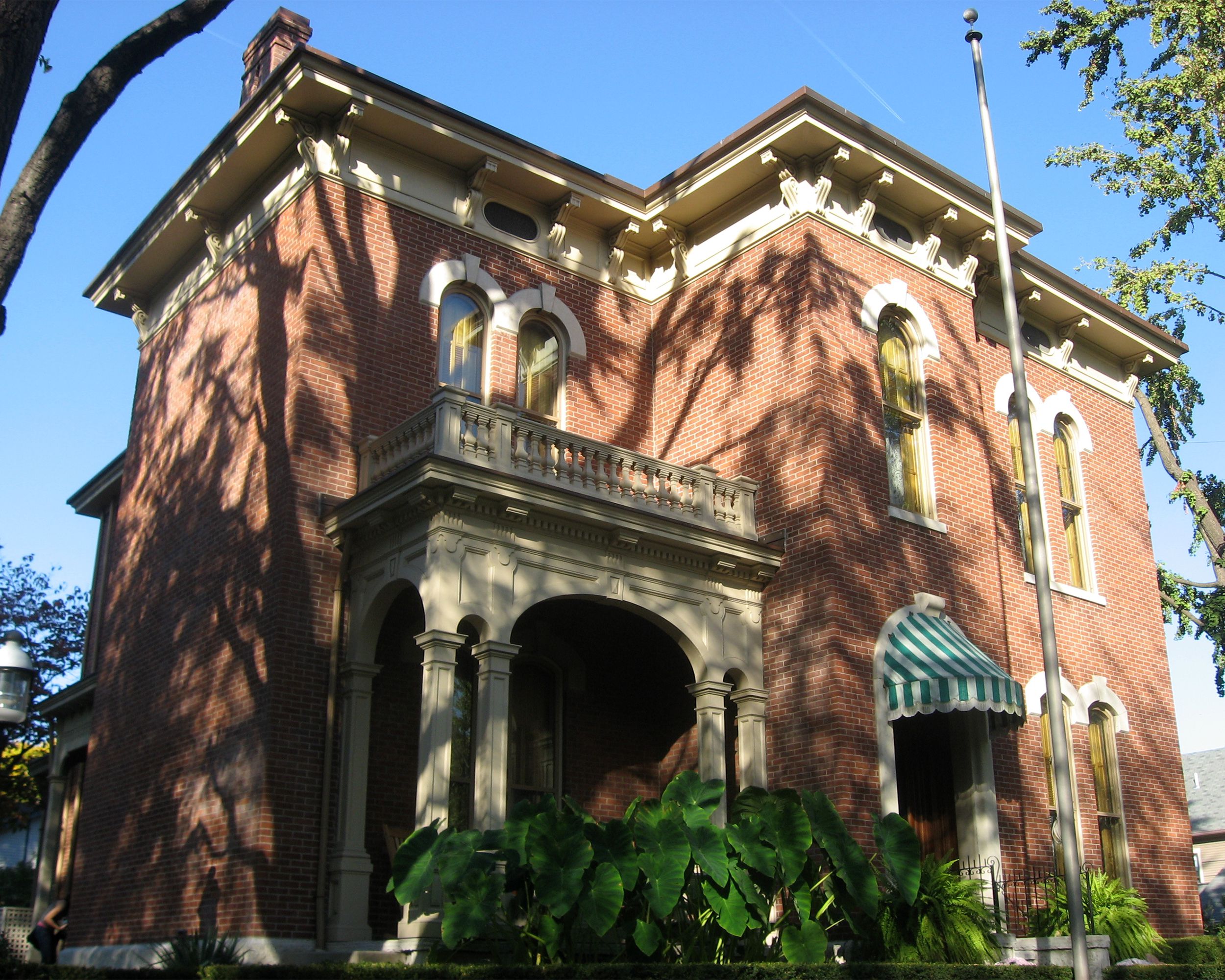 Front exterior of James Whitcomb Riley House, Indianapolis, red bricks with white wooden accents, tree shadows dramatically over the home, on a sunny day in spring