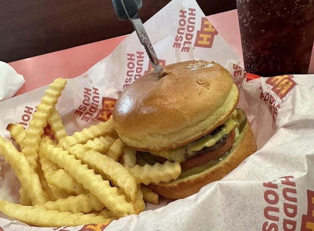 A cheeseburger with a knife stuck in the bun is served with crinkle-cut fries on Huddle House branded paper. A soft drink in a plastic cup is visible in the background.