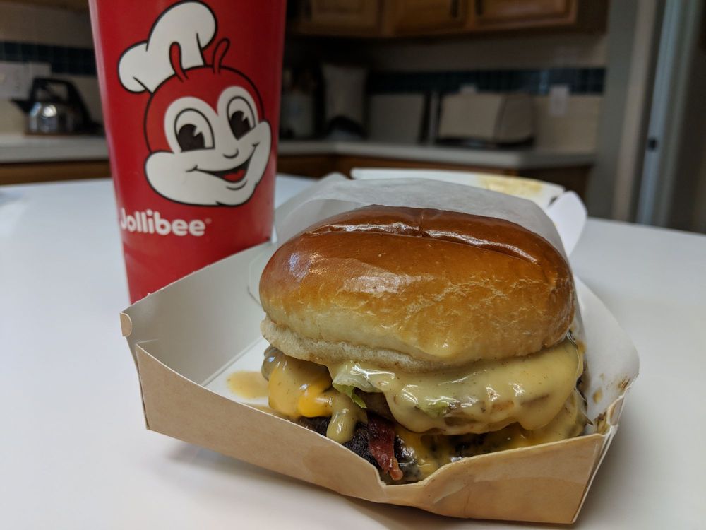 A cheeseburger with melted cheese and bacon sits in a cardboard box next to a large red Jollibee cup, all placed on a white kitchen counter with blurred cabinets in the background.