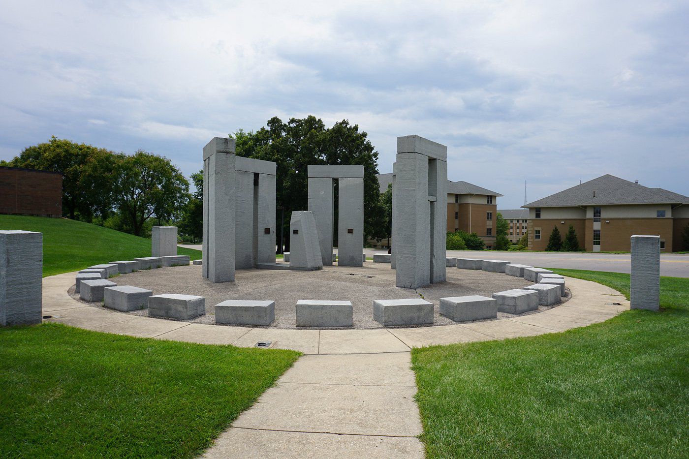 Missouri S&T Stonehenge, Rolla, Missouri, made out of granite surrounded by a round walkway and grass, trees and buildings in the background on a summer day