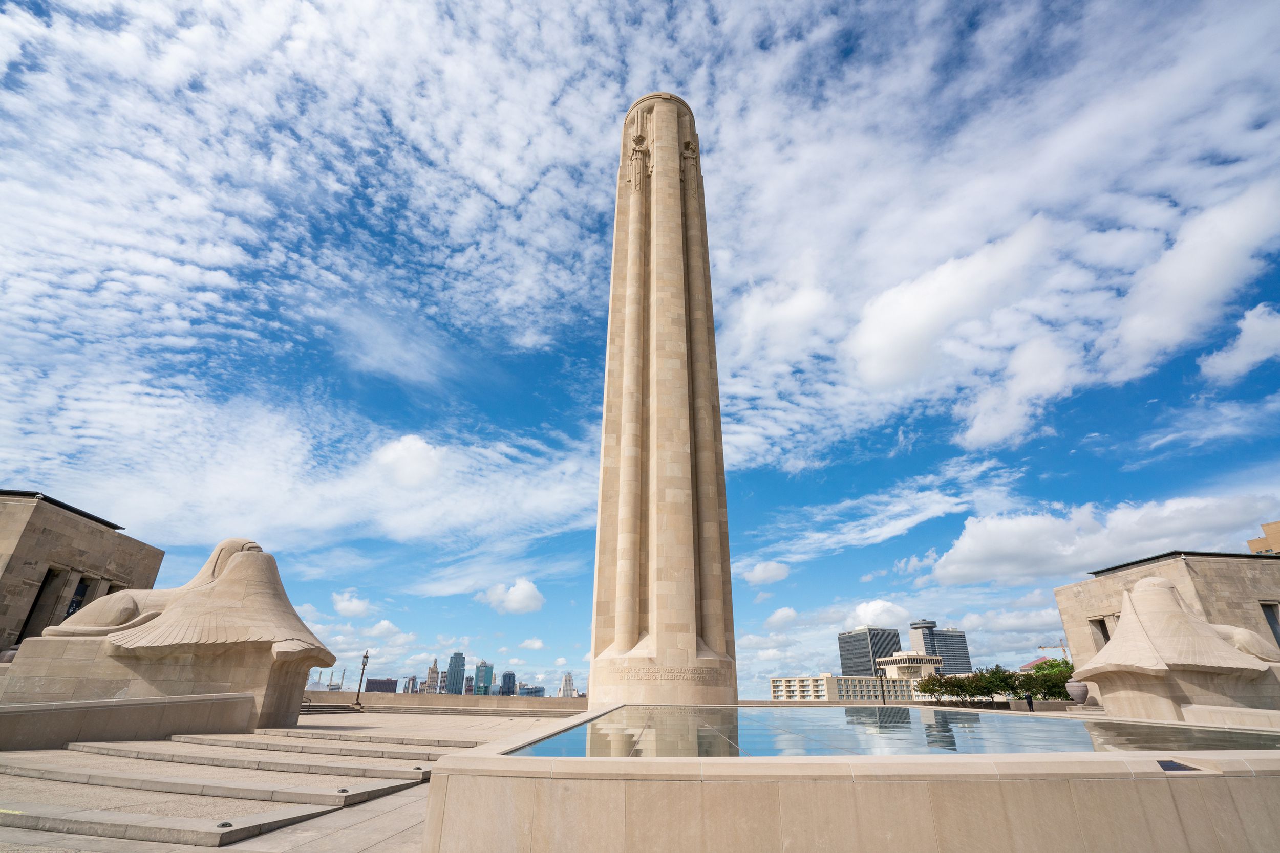 National World War I Museum & Memorial, Kansas City, Missouri, large slim tall monument surrounded by pools of water reflecting it, beige stone, a dramatic blue sky with white clouds with cityscape in the distance