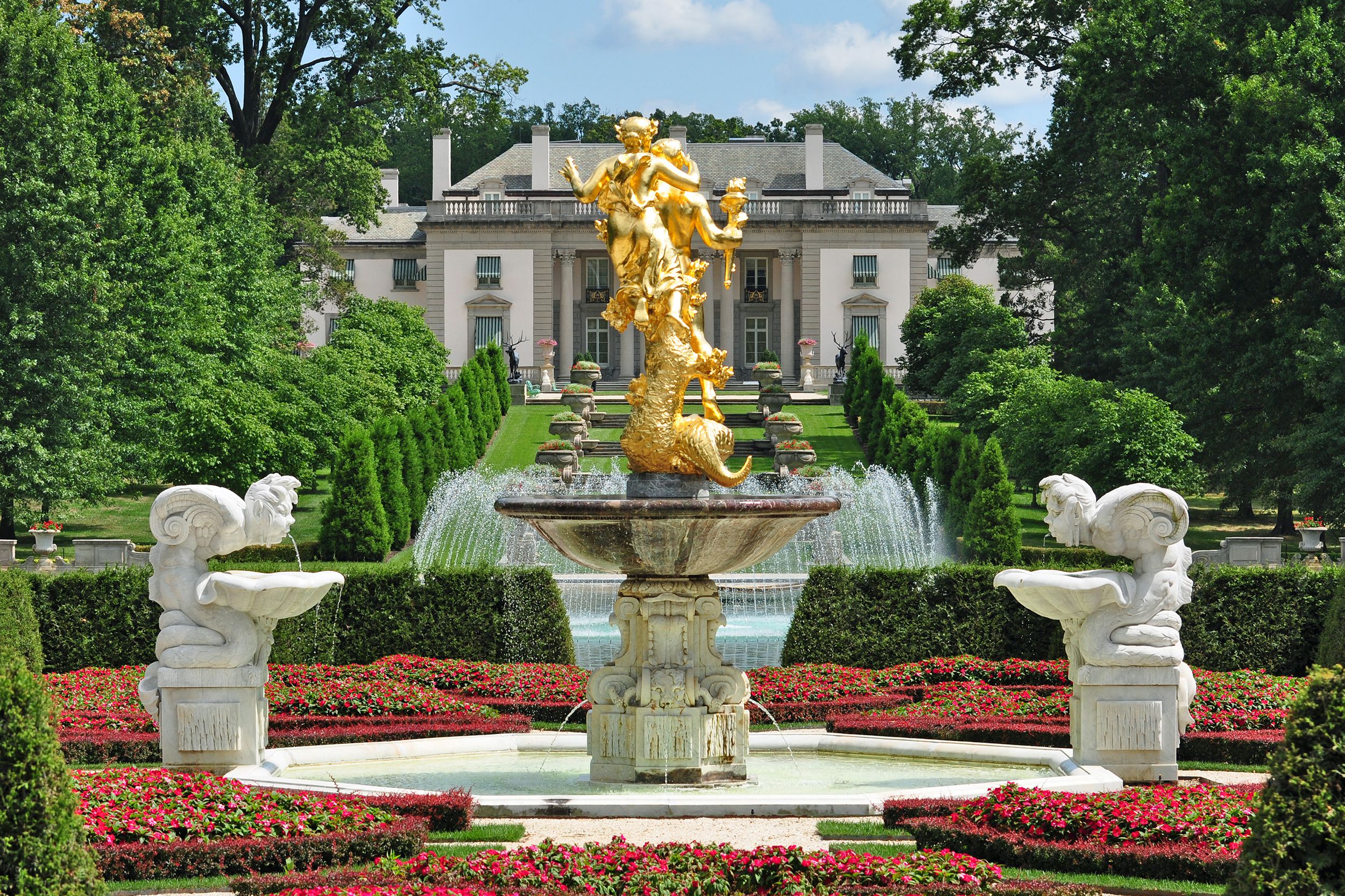 Nemours Mansion & Gardens, Wilmington, Delaware, gold statue of a fountain in the foreground surrounded by red flowers with garden going toward house in the background, on a sunny day during summer