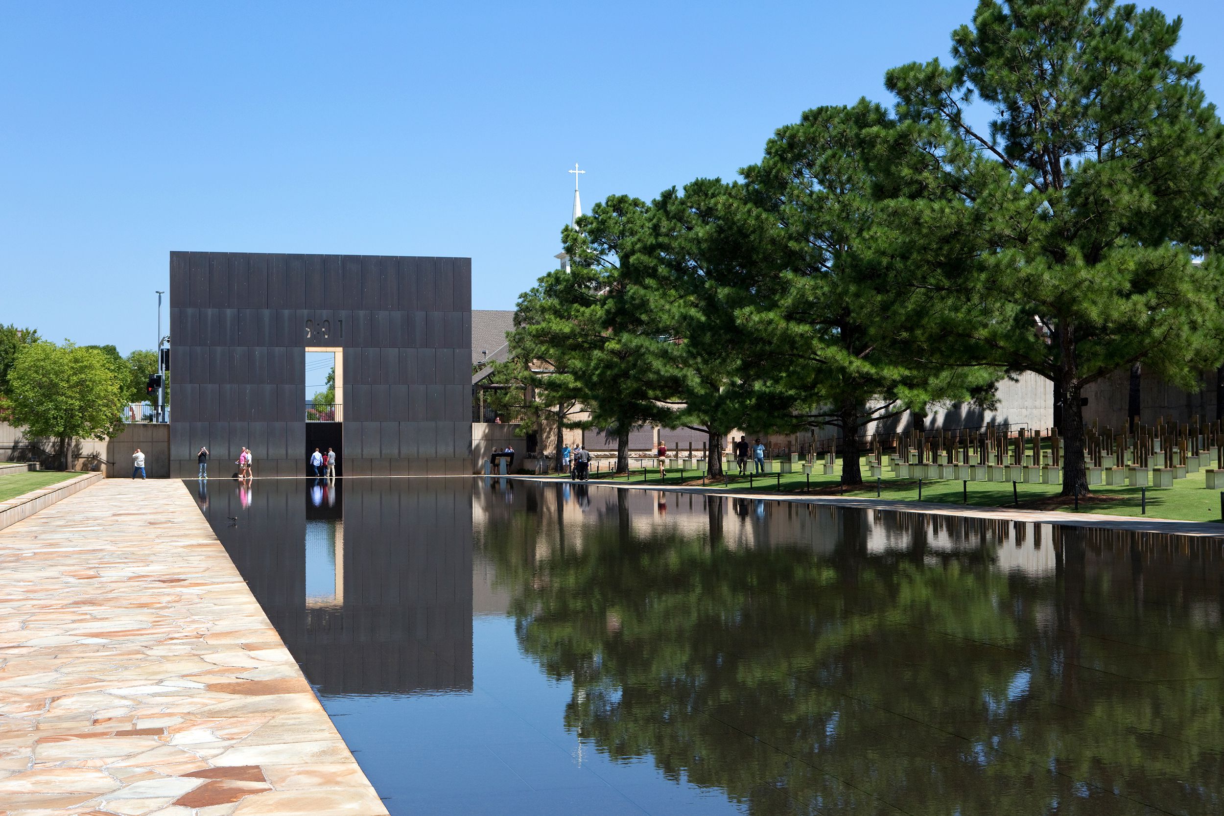 Oklahoma City National Memorial & Museum, Oklahoma City, pool in the foreground with stone walkway on the left and square grey granite memorial in the background, on a sunny day with a clear blue sky