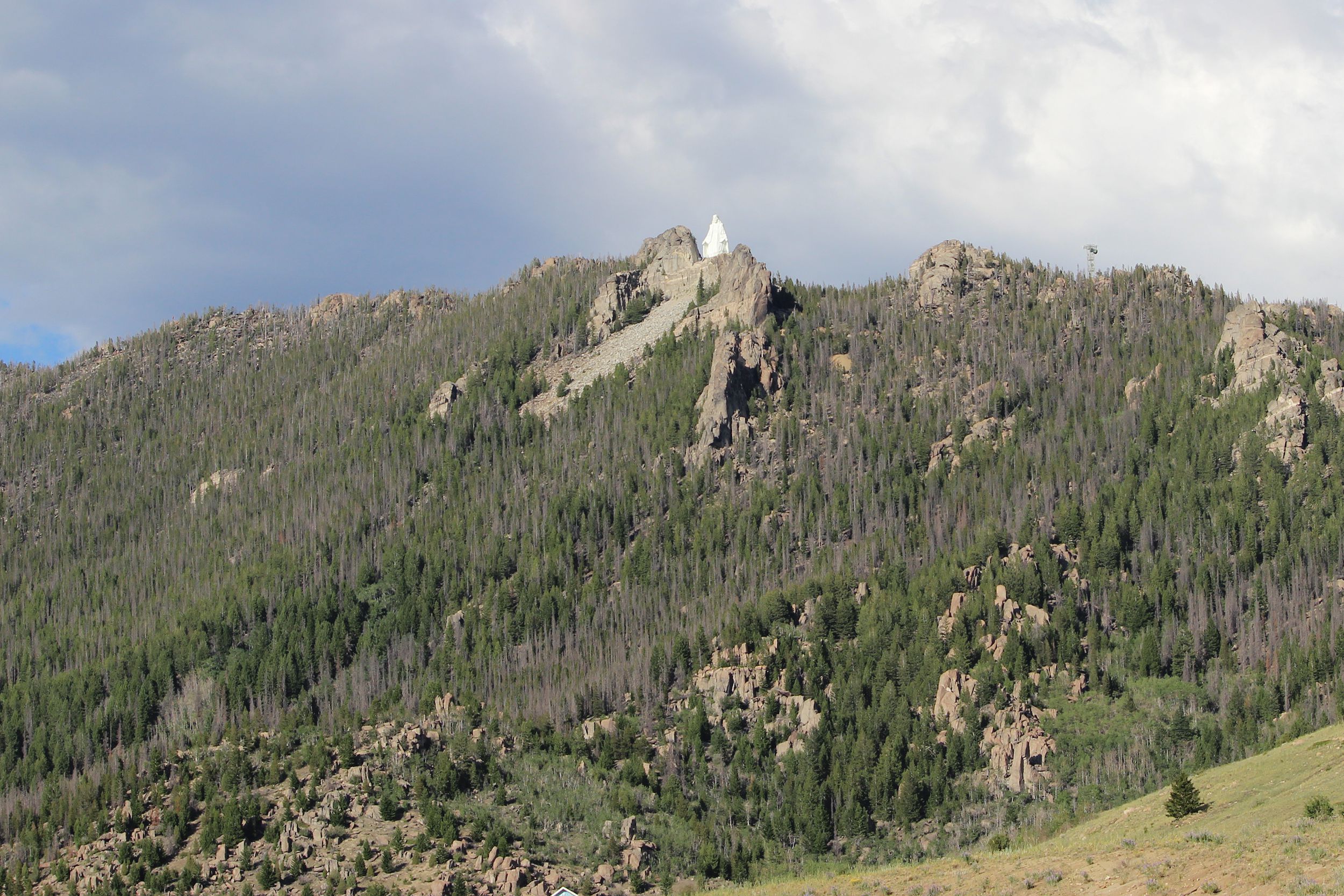 Our Lady of the Rockies, Butte, Montana, tiny white statue on top of expansive mountains, grey and white clouds in the background on a sunny day during summer