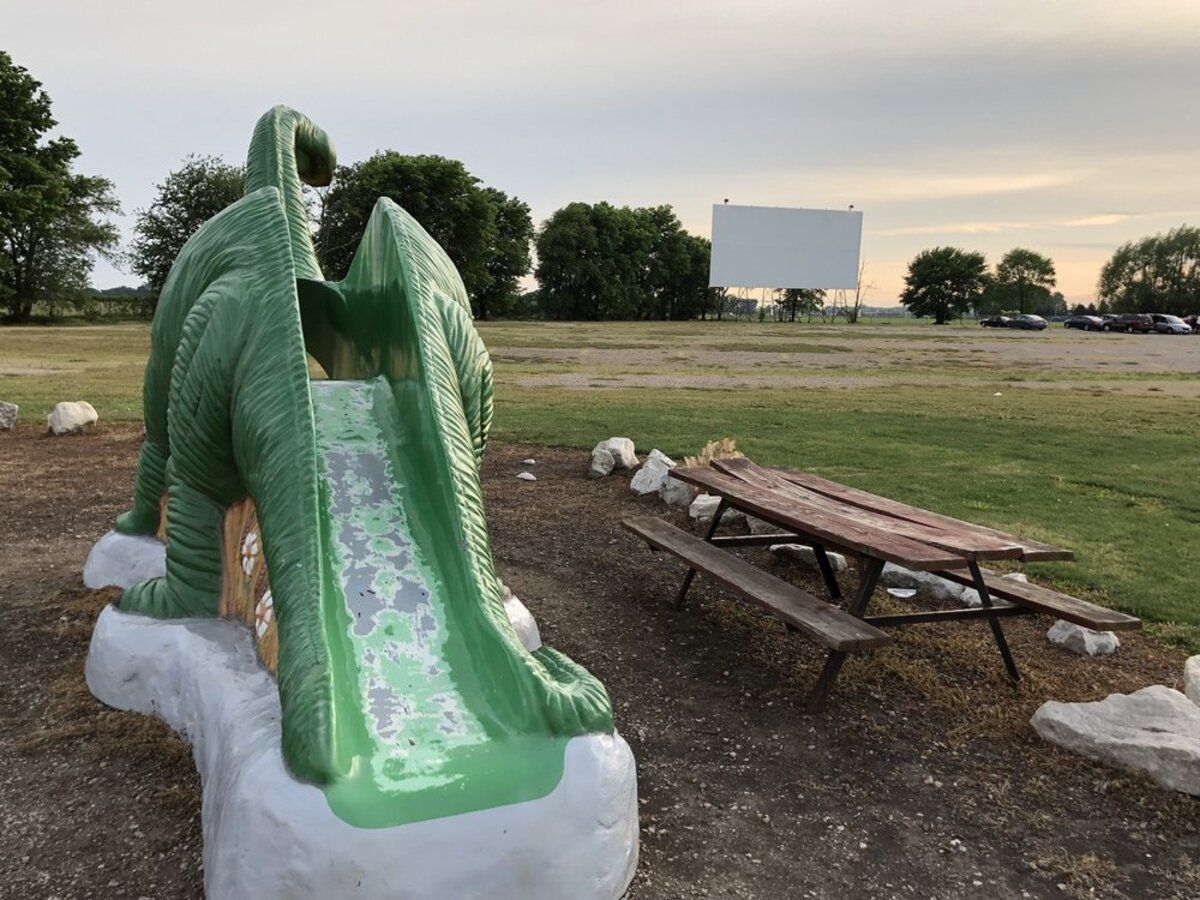 Outside movie screen of Route 66 Drive-in Theater, Springfield, Illinois, brontosaurus slide in the left and seating table in the right foreground, field and movie screen in the background, during early sunset