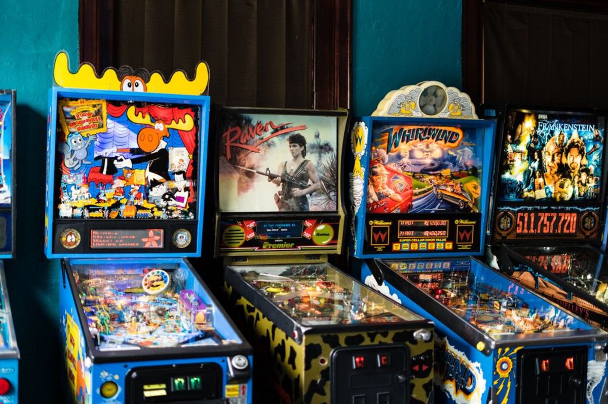 Row of pinball machines along a blue-green wall with brown shades at Route 66 Arcade Museum, McLean, Illinois