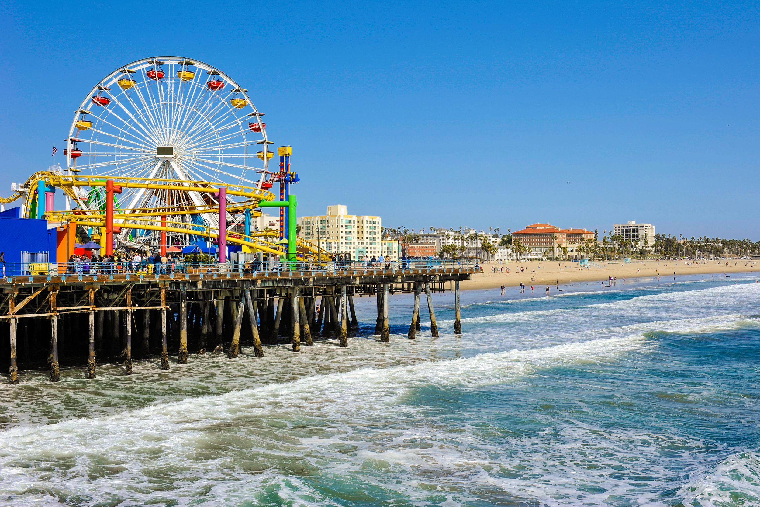 Santa Monica Pier, Santa Monica, California, pier on the left with ferris wheel and rides, ocean in the foreground, beach and buildings in the background on a sunny day