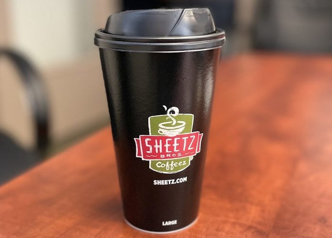 A black Sheetz Bros. Coffee cup with a plastic lid sits on a wooden table. The cup features the Sheetz logo, website, and "large" written at the bottom.