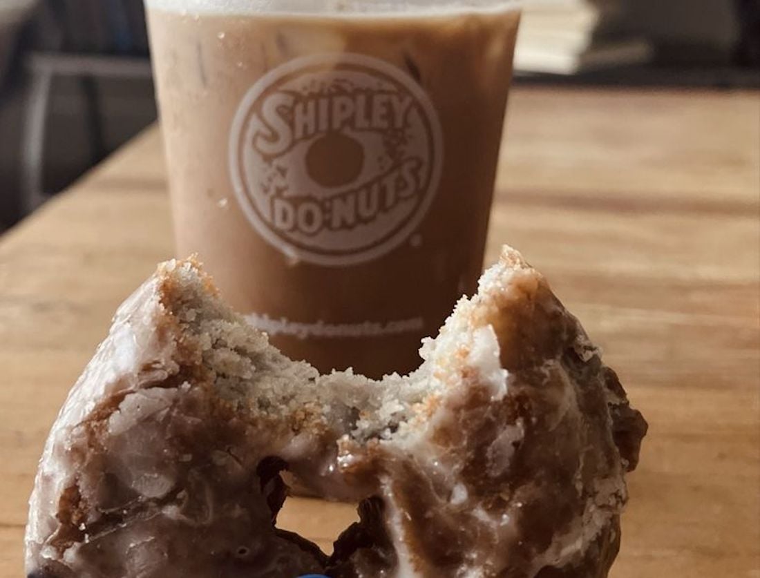 A close-up of a glazed donut with a bite taken out, sitting in front of an iced coffee in a clear Shipley Do-Nuts cup on a wooden table.
