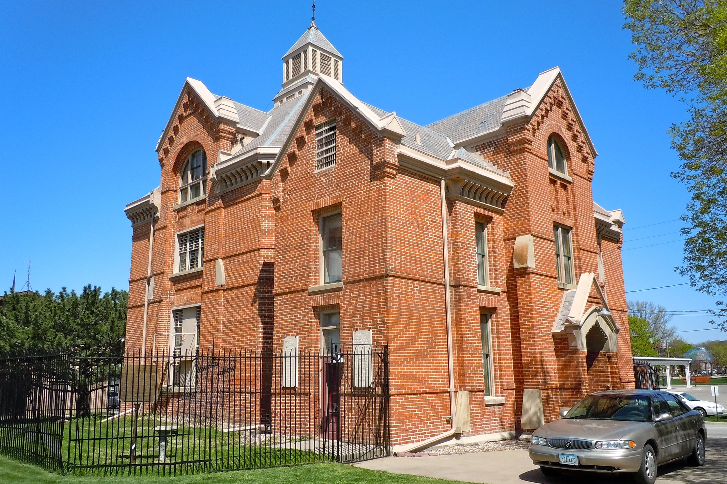 Front and side red brick exterior of Squirrel Cage Jail Museum, Council Bluffs, Iowa, surrounded by a black fence and grass, trees in the background on a sunny day during summer