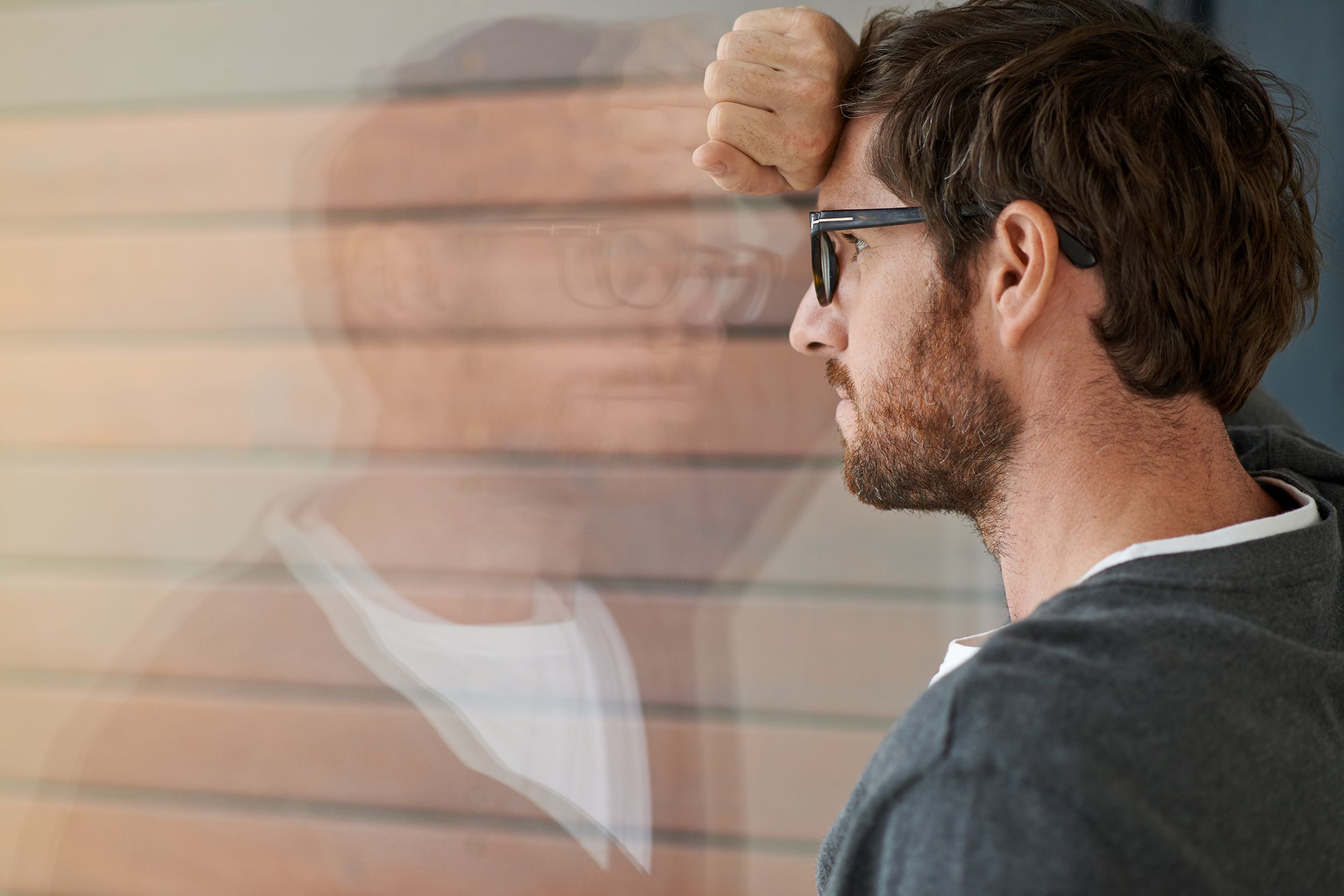 Worrying young man leaning his head against glass of a window, looking out of it, with faded reflection of him in the glass and wall in the background