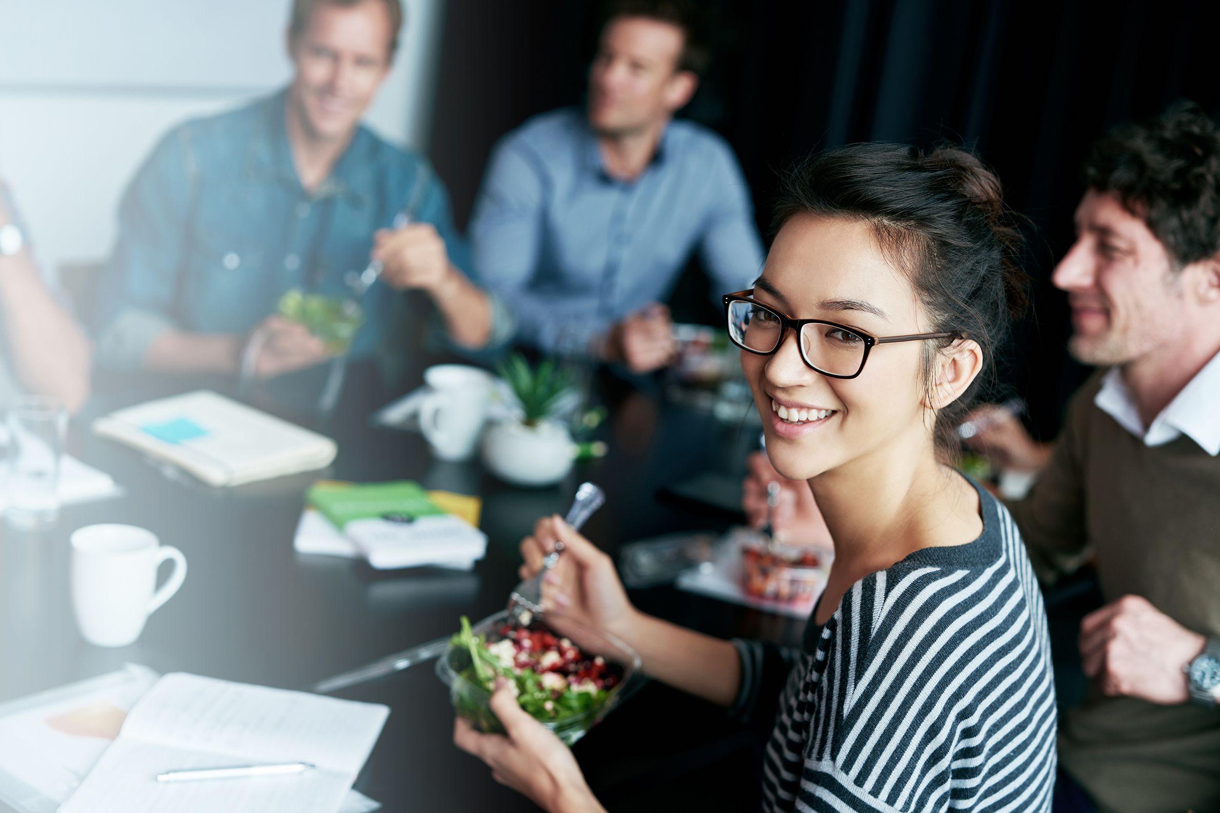 Young woman smiling towards the camera while holding a bowl of salad, eating with a fork, around a boardroom table with other work colleagues sitting around it, blurred and light coming from left side