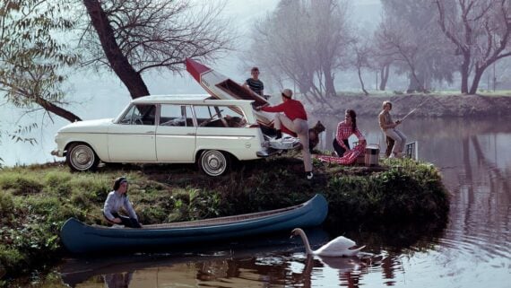 A family enjoys a day by a lake with a white vintage car and an open trunk. Some fish by the water, one person sits in a blue canoe, and a swan swims nearby. Bare trees and a misty background set the scene.