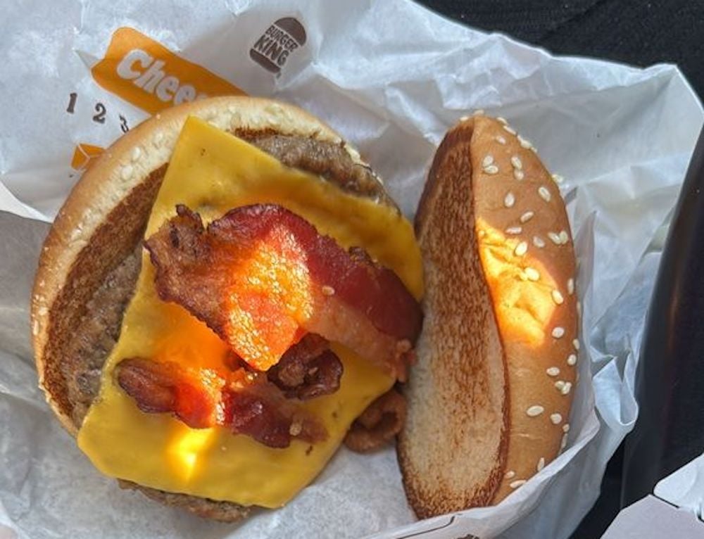 A cheeseburger with bacon strips, a slice of cheddar cheese, and a beef patty on a sesame seed bun, partially unwrapped from Burger King packaging.