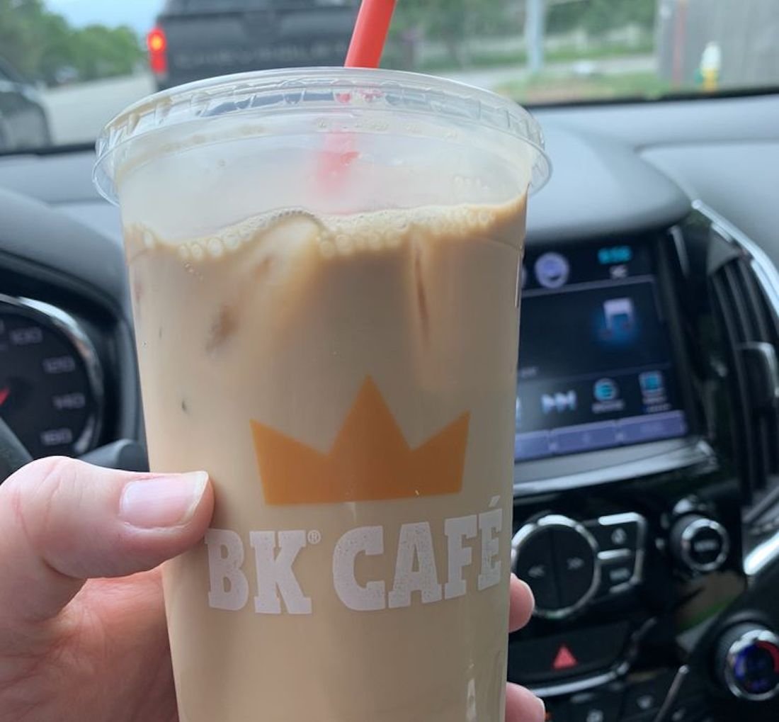 A hand holds a large iced coffee in a plastic cup with a red straw and "BK Café" logo, inside a car with the dashboard and radio visible in the background.