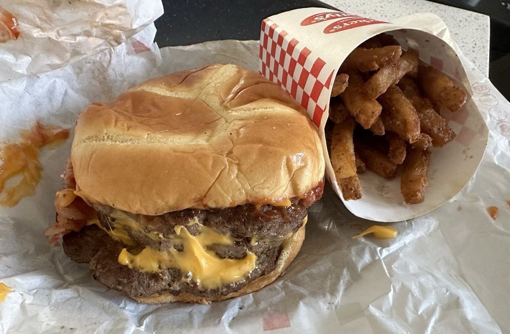 A cheeseburger with two beef patties and melted cheese sits on a wrapper next to a container of seasoned curly fries.