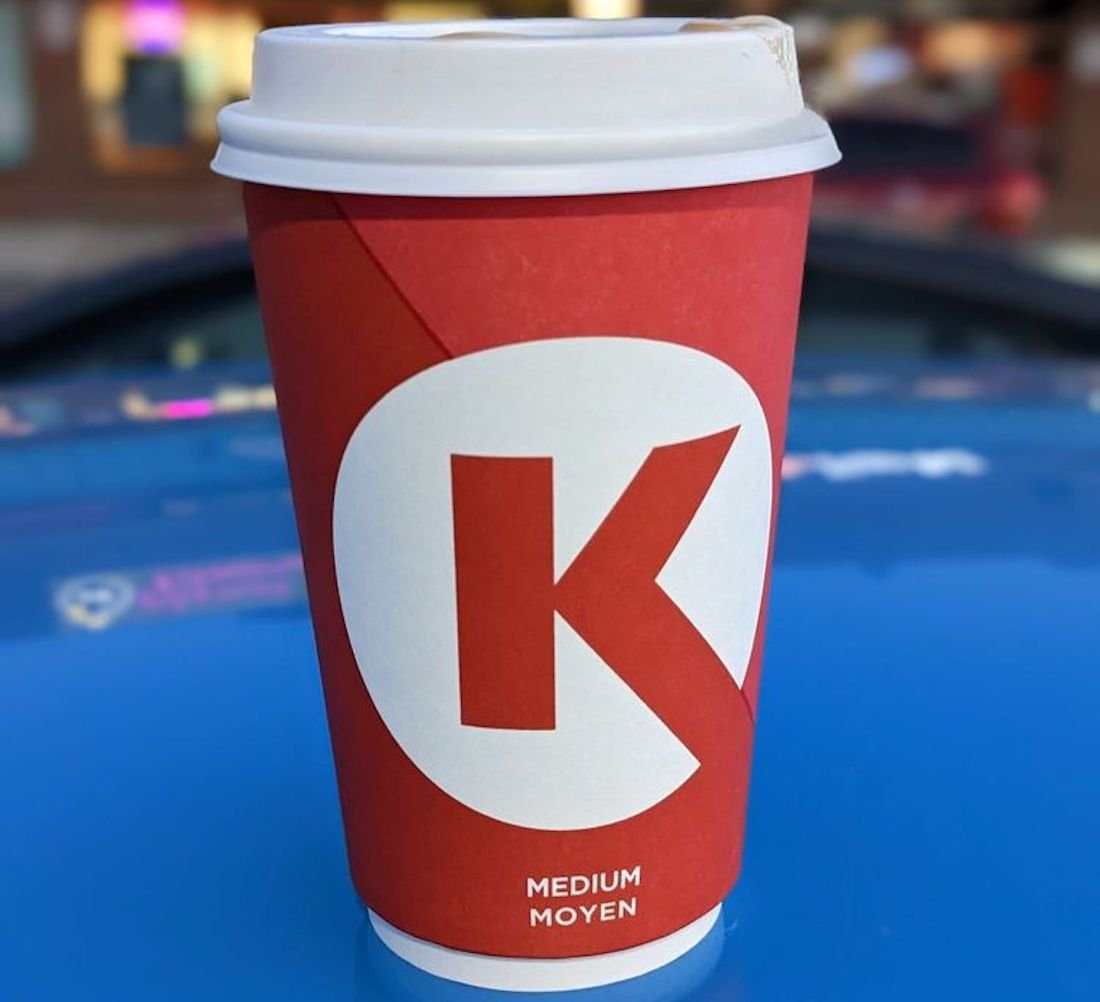 A red takeout coffee cup with a large white "K" logo and the word "MEDIUM" in English and French, sitting on a blue surface with a blurred background.