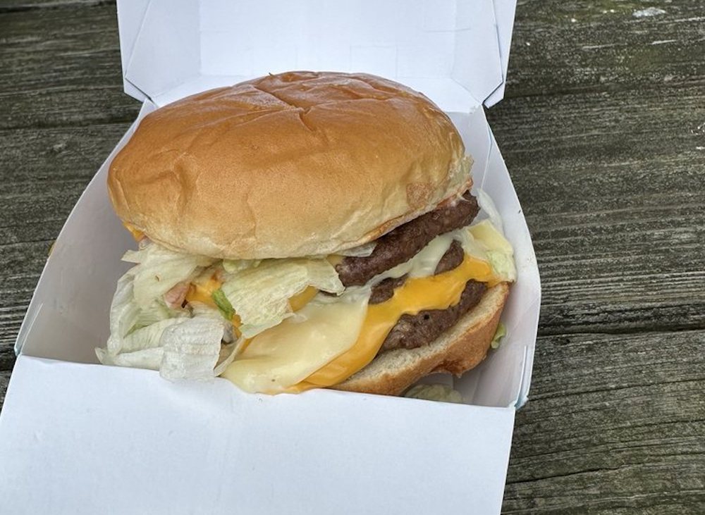 A cheeseburger with two beef patties, cheddar and Swiss cheese, lettuce, and sauce in a cardboard takeout box on a wooden surface.