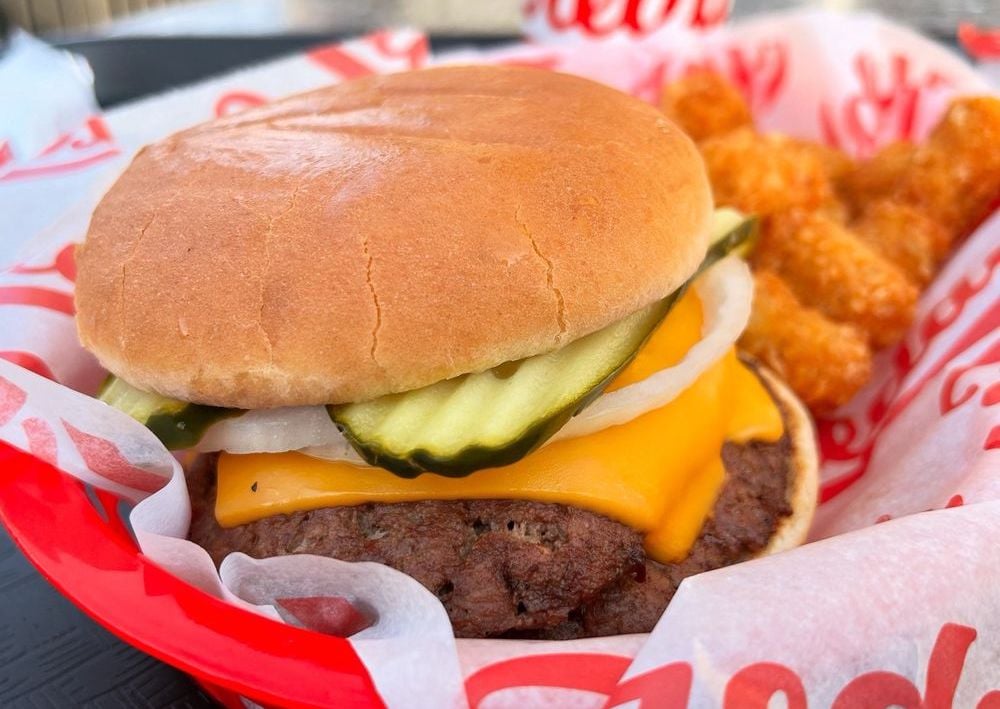 A cheeseburger with pickles, onions, and cheddar cheese in a soft bun sits in a red basket beside crispy tater tots, on white paper with red text.