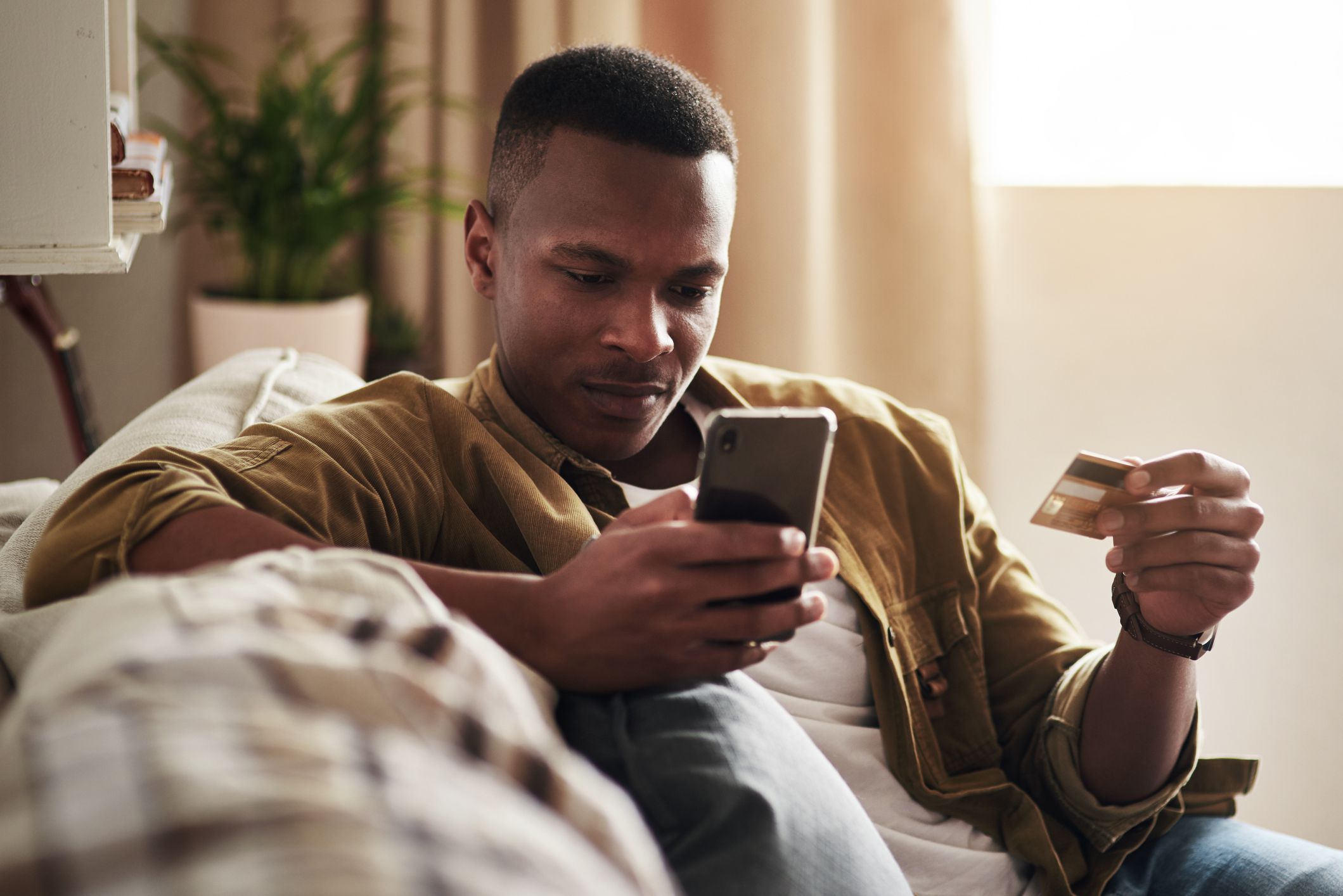 Cropped shot of a handsome young man using a smartphone and a credit card to shop online while sitting on his couch at home