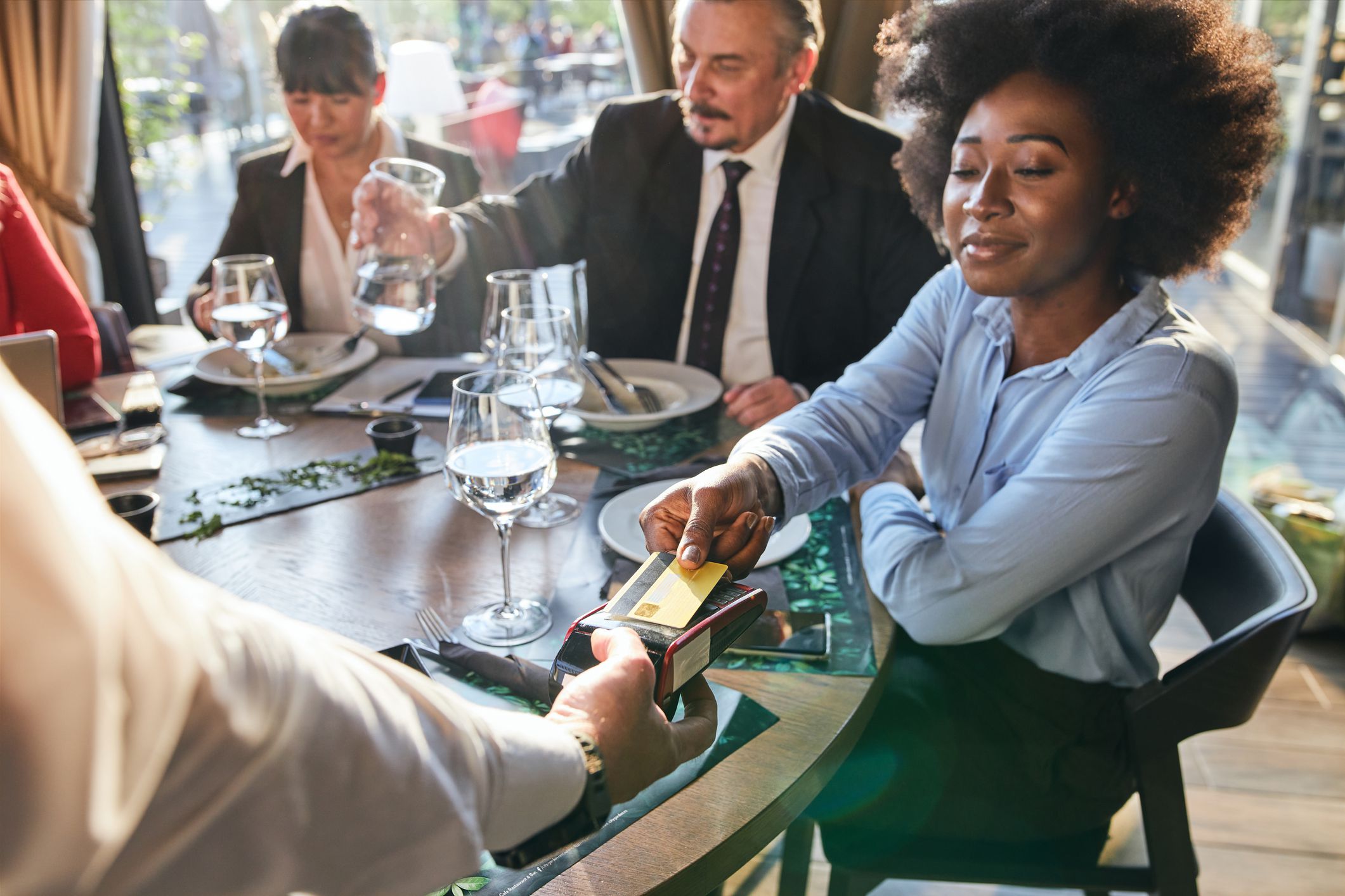 A diverse group of businesspeople enjoying lunch at a restaurant, and an African-American woman paying with a credit card via contactless payment