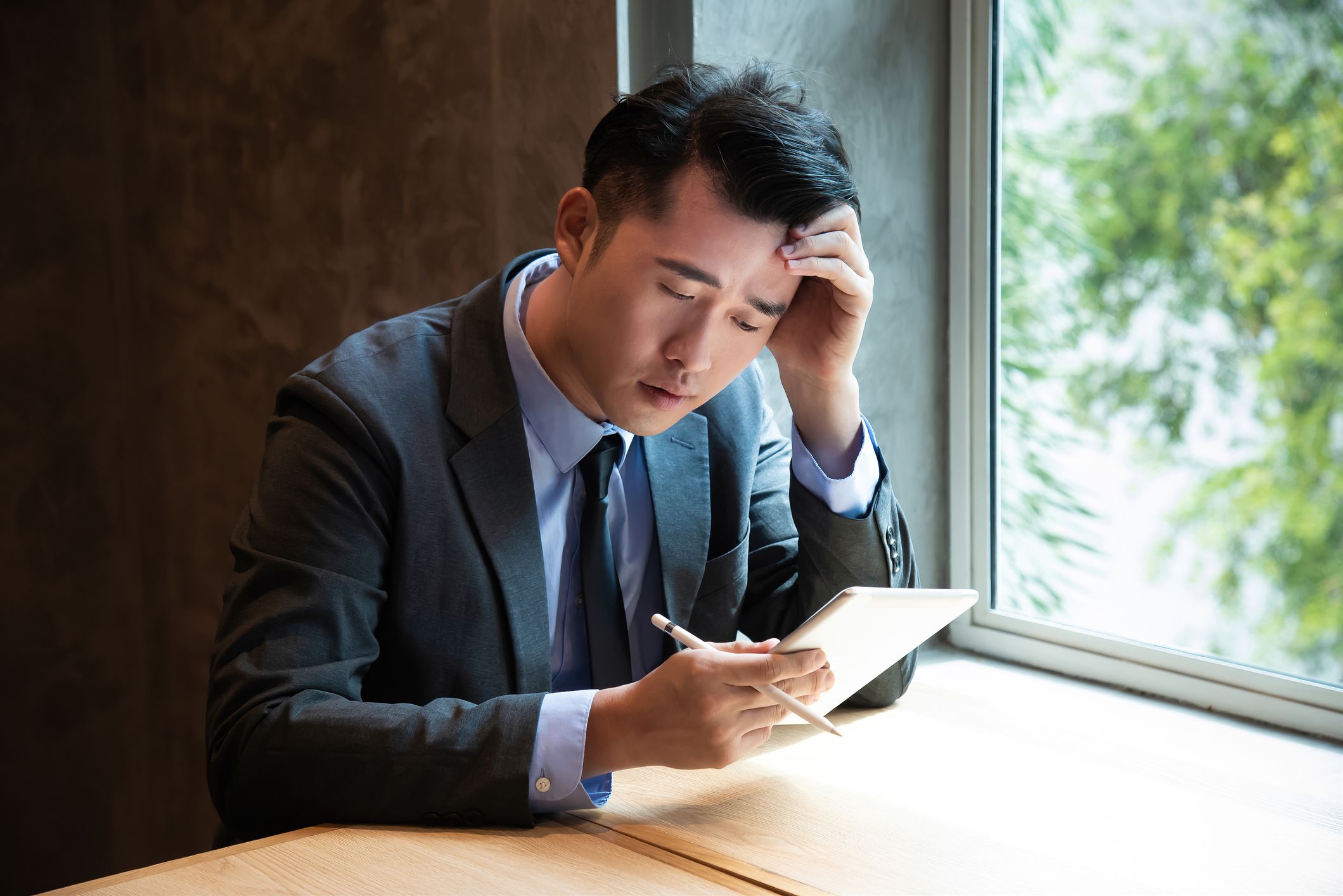 Desperate Asian businessman sitting hopelessly in office received bad news from broker about his bad stock market investments. Businessman receiving bad stock market investment report with digital tablet while sitting at office desk.