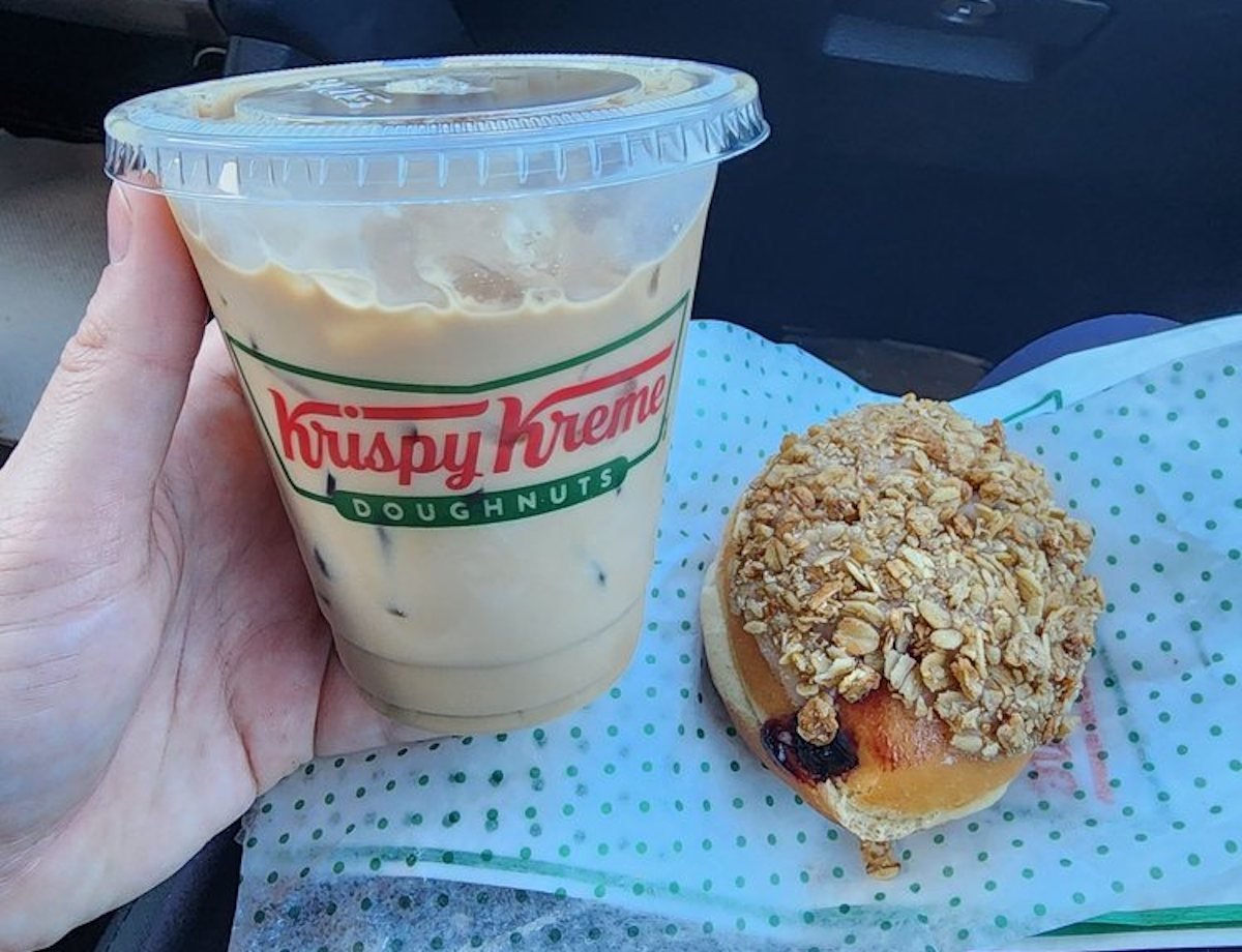 A hand holds an iced coffee in a Krispy Kreme cup next to a doughnut topped with oat crumble, both resting on a Krispy Kreme wrapper.