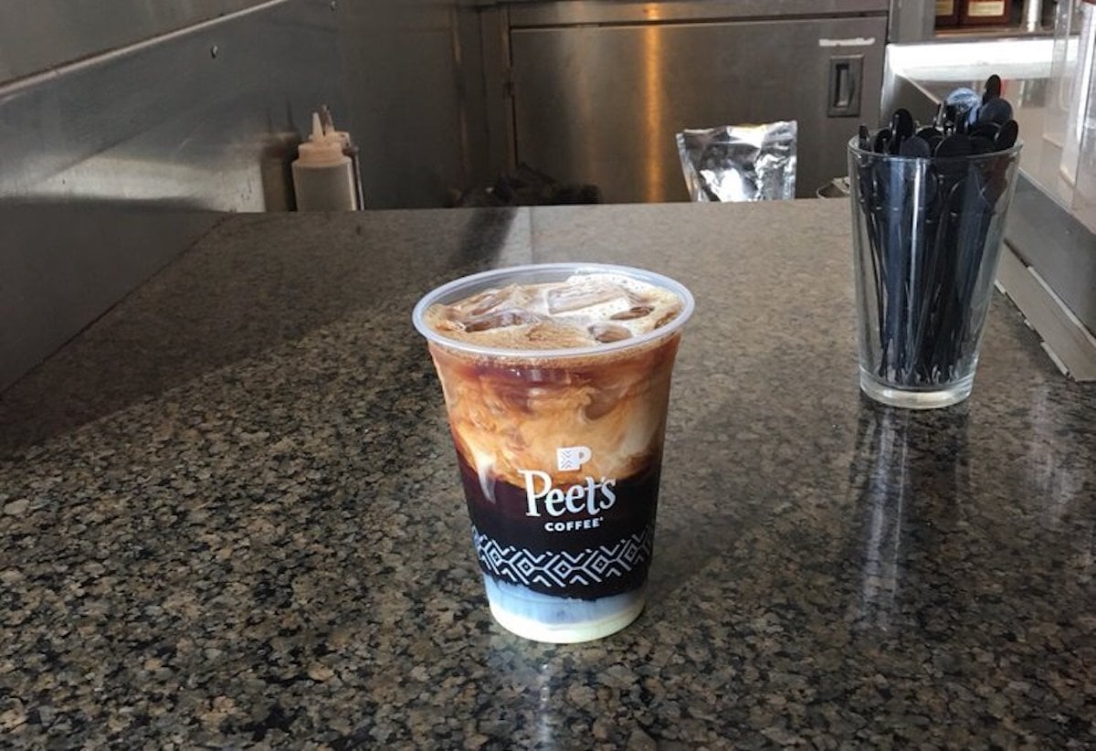 A plastic cup of iced coffee with cream, labeled "Peet's Coffee," sits on a granite counter. In the background are a container of black straws and part of a stainless steel kitchen area.
