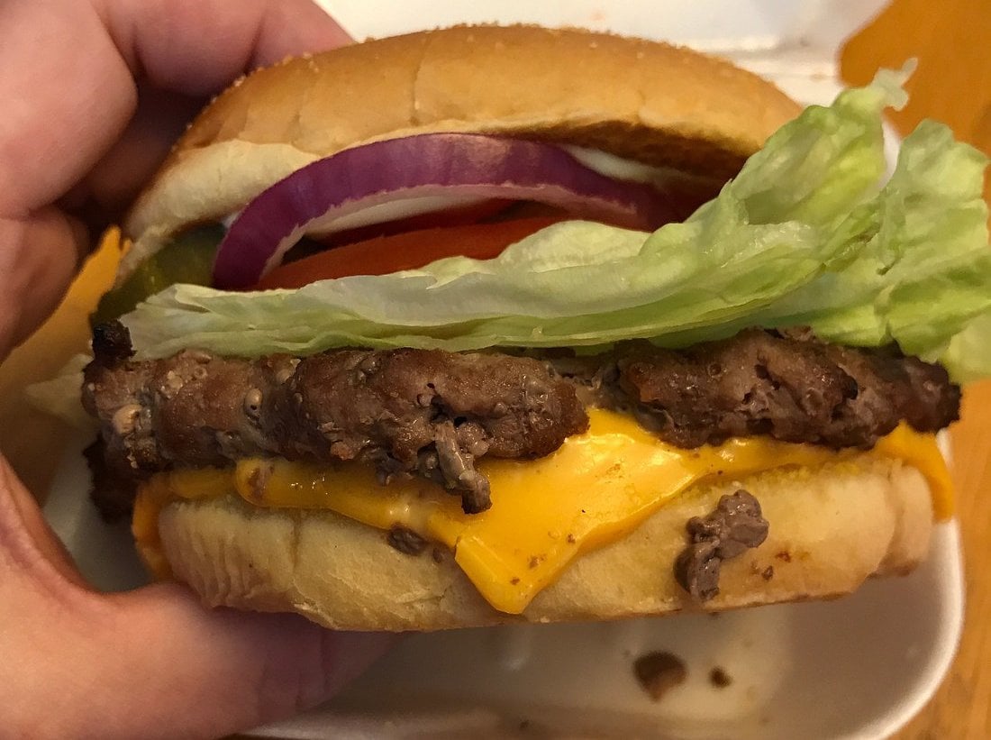A close-up of a hand holding a cheeseburger with lettuce, tomato, pickles, red onion, cheddar cheese, and a beef patty in a sandwich bun.