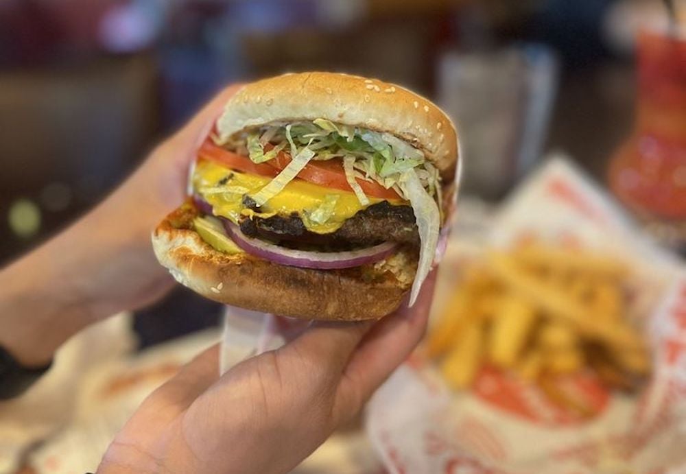 A person holds a cheeseburger with lettuce, tomato, onions, pickles, and melted cheese in a sesame seed bun. In the background, there is a tray with French fries.