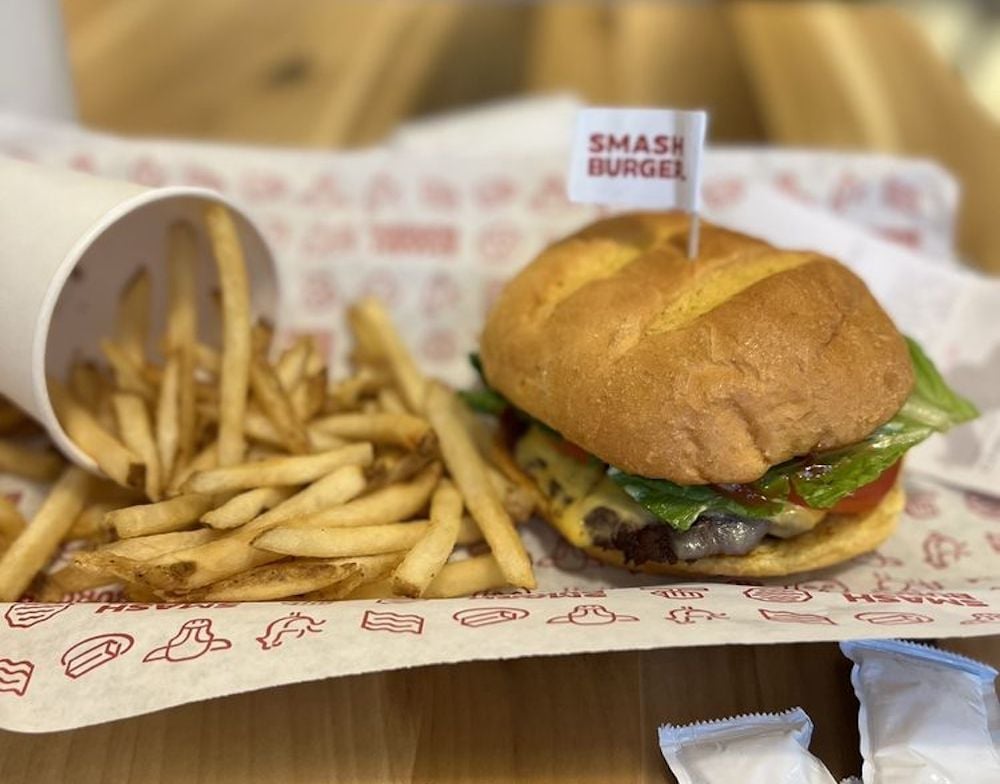 A cheeseburger with lettuce and tomato, topped with a small "Smash Burger" flag, sits next to a pile of French fries spilled from a paper cup on a tray lined with branded paper.