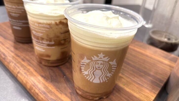 Three Starbucks cold drinks topped with foam are placed on a wooden tray. The drinks are in clear plastic cups displaying the Starbucks logo. The focus is on the front cup, with two others blurred in the background.