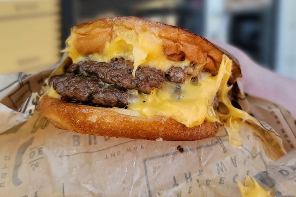 Close-up of a Wayback cheeseburger with two beef patties and melted cheese, served in a soft bun with cheese oozing out, resting on branded parchment paper.
